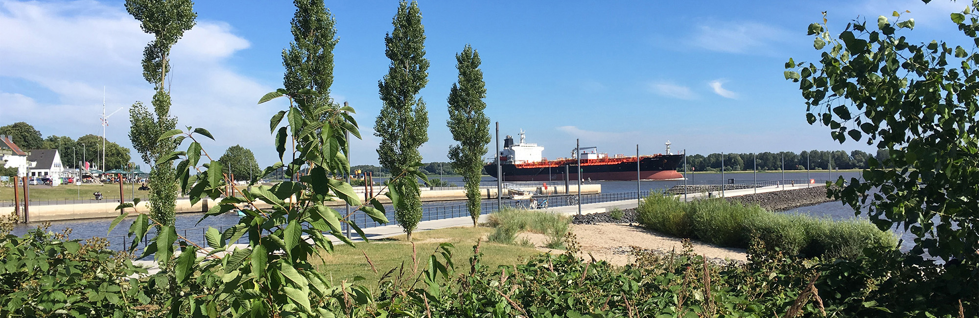 Immobilienmakler Wedel - Das von Hohorst & Schwier erstellte Bild zeigt den neuen Schulauer Hafen in Wedel. Auf der Elbe sieht man ein vorbeifahrendes Frachtschiff während im Vordergrund der Hafen und die Promenade zu erkennen ist. Die Aufnahme wurde an einem sonnigen Tag gemacht.